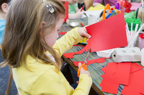 Little girl using scissors to cut shapes from a red piece of paper. she is in a nursery class with other students.